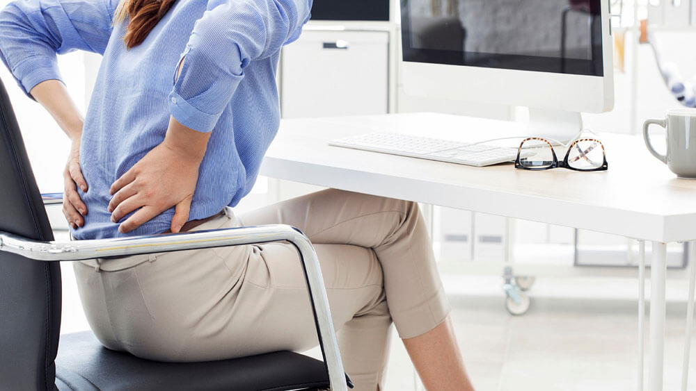 Woman sitting at desk in office chair holding her lower back as if in pain
