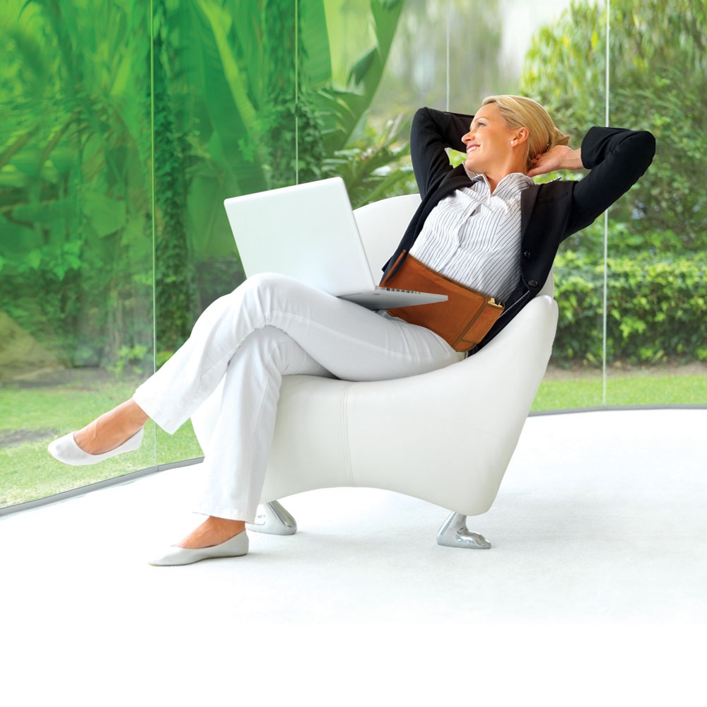 Woman sitting in chair inside room with large windows looking out into a lush green landscape. She has her hands behind her head, relaxing, with a laptop in her lap and a Richway Bio-Belt around her waist.