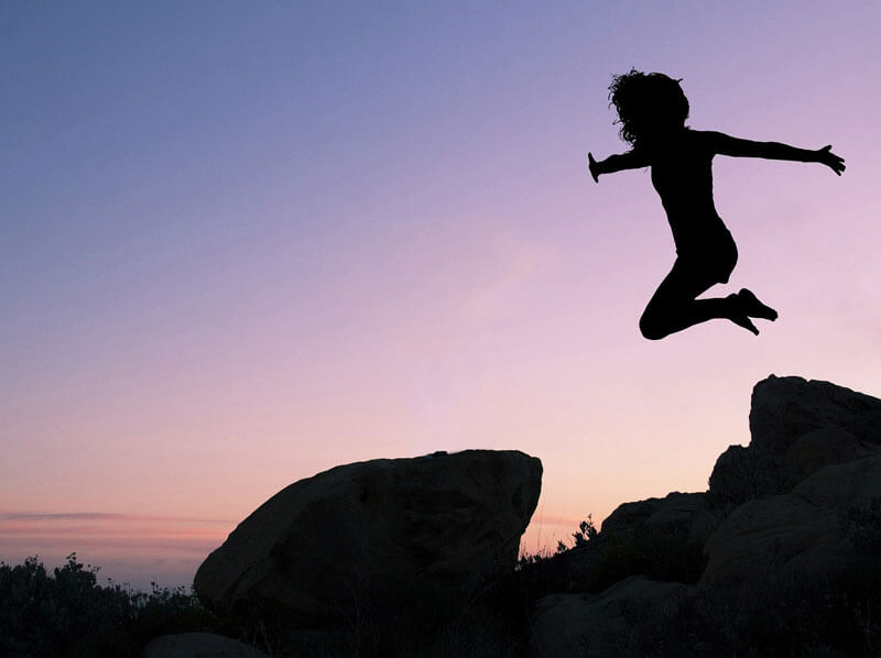 woman silhouette jumping off rocks with energy at the beach against a sunset background.