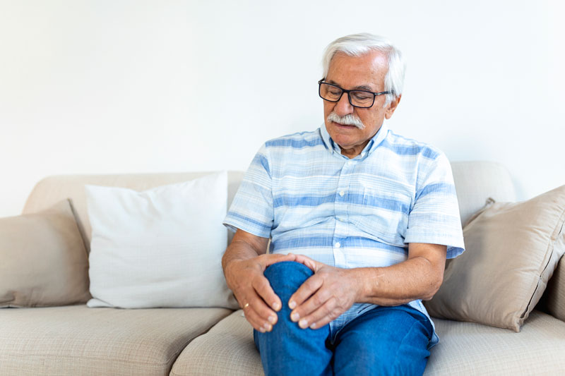 Older man sitting on couch clutching his knee as if in pain.