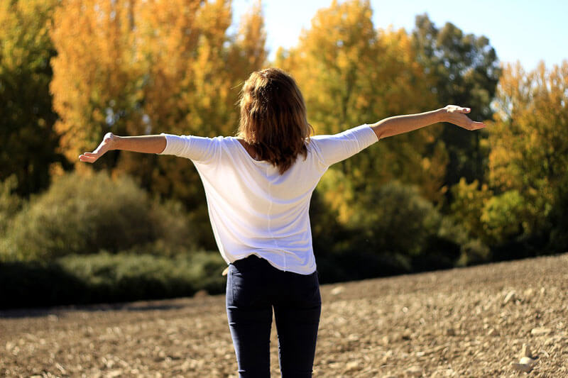 woman with her back to the camera out in nature, standing in front of a forest with her arms above her head in jubilation.