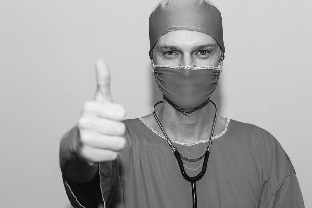 Doctor with scrubs and a surgical mask on holding his right arm out with a thumbs up
