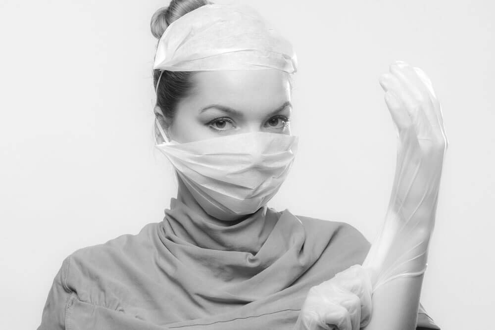 Female doctor with one arm raised putting on surgical gloves. Doctor has surgical mask on and scrubs.
