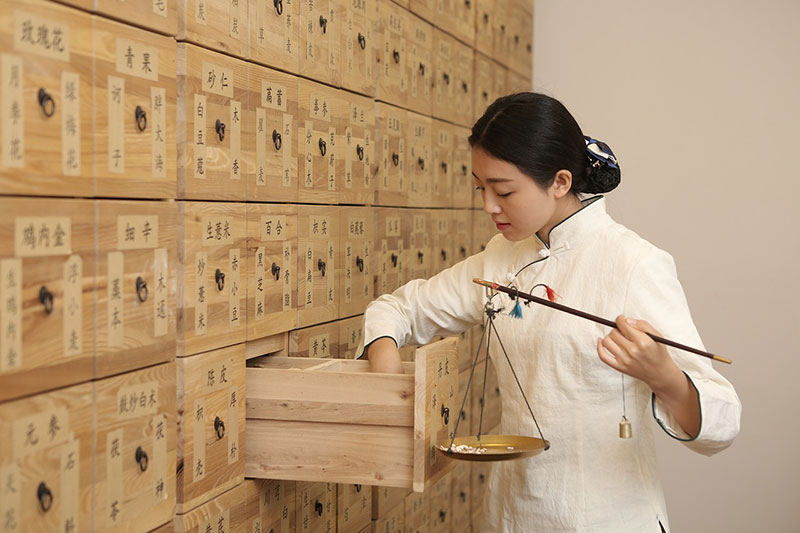 Chinese doctor next to a wall of drawers that hold herbs weighing out herbs.
